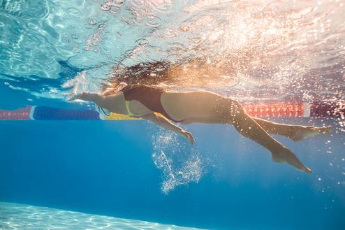 Unterwasseraufnahme einer Schwimmerin in einem Bikini, die kräftig durch das Wasser stößt, mit bunten Schwimmbahnmarkierungen.